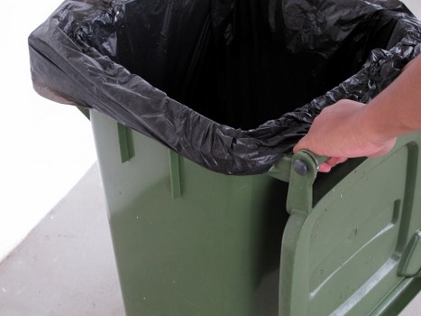 A close-up view of a green plastic rubbish bin with a black plastic trash bag inside, partially visible at the top. A person's hand is gripping the side of the bin's lid, which is slightly open, revealing the bag's edges folded over the rim. The bin is positioned on a light-colored pavement or driveway surface, with a plain background and no other objects visible. The scene is well-lit, suggesting natural daylight, and the setting appears to be outdoors, possibly in an urban or residential area within Pimlico. Commercial Waste Removal Pimlico provides rubbish collection services, and this image depicts a typical waste collection or disposal container ready for collection or disposal processes.