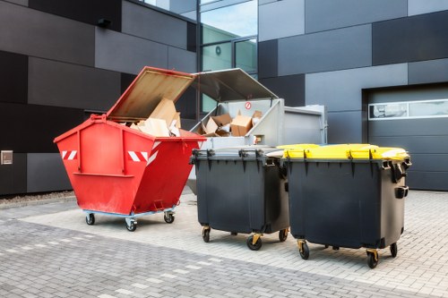 A row of three large wheelie bins is positioned on a paved outdoor area beside a modern building with dark grey and black panels. The first bin, which is red, has its lid open, revealing a collection of cardboard boxes and packaging materials inside, with some boxes leaning against the sides. Next to it are two black bins with yellow lids, both of which are closed and appear to be in good condition. The scene suggests waste collection or rubbish removal activity, typical of commercial waste services in Pimlico. The environment is clean, and the bins are situated on a concrete or stone surface, with minimal surrounding clutter, emphasizing their purpose for rubbish disposal. The modern building environment and the arrangement of waste containers reflect common street-front or commercial premises in the area, supporting services like those provided by Commercial Waste Removal Pimlico.