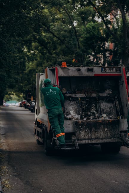 A worker in a yellow safety vest and red shirt is operating waste collection machinery at the rear of a large red garbage truck on a street. The truck, branded with identifiable safety markings and li
