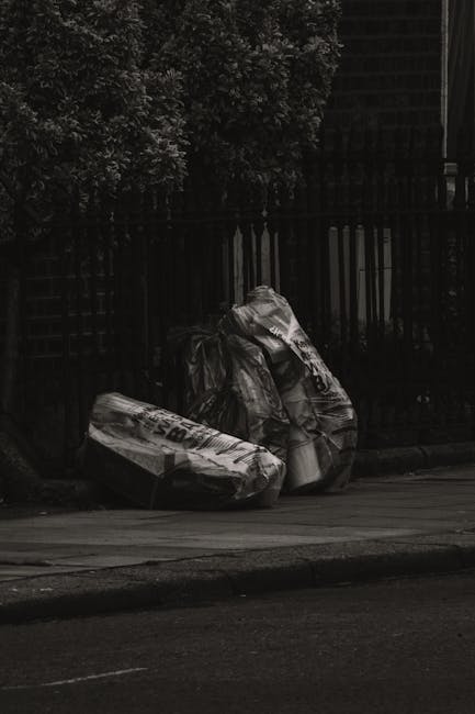 Two large black garbage bags, made of thick plastic with a slightly crinkled texture, are placed on the pavement next to a black metal fence with vertical bars. The bags appear to be filled with waste