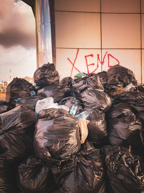 Two large black garbage bags, made of thick plastic with a slightly crinkled texture, are placed on the pavement next to a black metal fence with vertical bars. The bags appear to be filled with waste